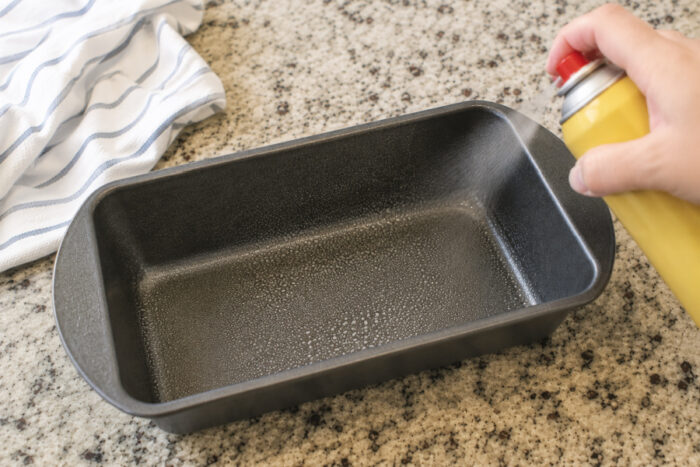 Greasing a loaf pan with nonstick cooking spray for moist pumpkin bread.