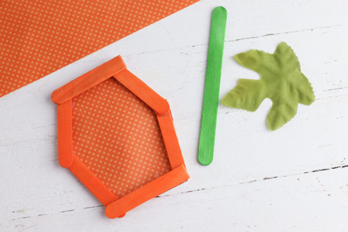 Orange scrapbook paper added behind the popsicle stick pumpkin frame with a green stick and faux leaf nearby
