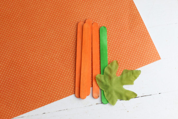 Orange and green popsicle sticks, orange scrapbook paper, and a faux leaf for a popsicle stick pumpkin craft