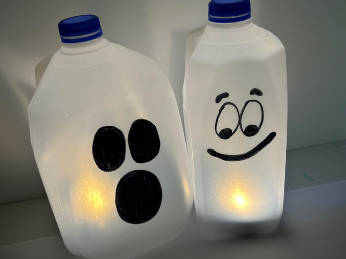 Two glowing milk jug ghosts with different black ghost faces and flameless lights inside.