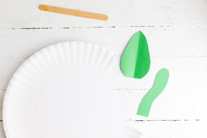 Supplies for making an apple paper plate craft including a paper plate, craft stick, and green construction paper leaf and caterpillar