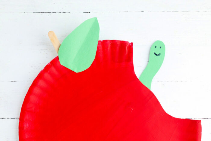 Completed apple paper plate craft with a green leaf, craft stick stem, and caterpillar peeking out of the bite