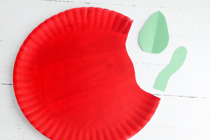 Red painted paper plate with green leaf and caterpillar cutouts ready to assemble into an apple craft